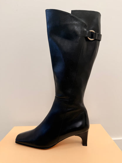 Black knee-high boot with a buckle on a beige surface and light gray background